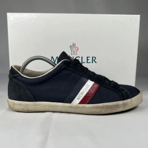 Moncler sneakers i marinblå mocka - Snygga sneakers från Moncler i marinblått tyg och mocka med klassiska ränder i rött, vitt och blått på sidan. Skorna har rund tå, platt sula och snörning framtill. Moncler-logga på plösen och sidan ger en exklusiv vibe. Perfekta för dig som gillar stilrena och sportiga skor.