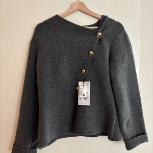 Grå ullkofta med guldknappar - Snygg grå kofta i ull med asymmetrisk knäppning och guldiga knappar framtill. Modellen har lång ärm och uppvikta muddar, perfekt för lager-på-lager. Enkel men ändå unik design som sticker ut.