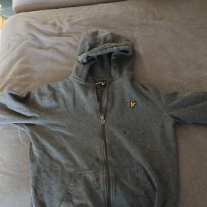 Grå hoodie från Lyle & Scott Junior - Mörkgrå hoodie från Lyle & Scott Junior med dragkedja framtill och klassisk gul logga på bröstet. Tröjan har huva, långa ärmar och två fickor framtill. Perfekt för chill dagar eller när du vill ha en enkel och stilren look.
