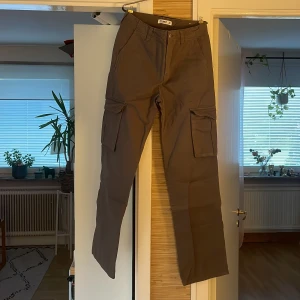 Grå cargobyxor med raka ben - Säljer ett par grå cargobyxor från Pull & Bear med raka ben och flera stora fickor på sidorna och baktill. Byxorna har klassisk cargostil och är tillverkade i slitstark bomull. Perfekta för dig som gillar praktiska och snygga byxor med streetkänsla.