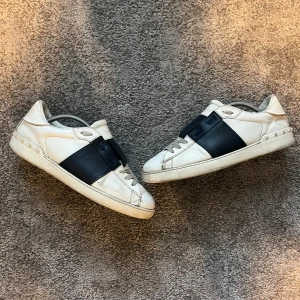 Valentino Open sneakers vit/navy skinn - Snygga Valentino Open sneakers i vitt skinn med bred svart läderdetalj över mitten. Klassisk rund tå, platt sula och diskret logga på plösen. Skorna har snörning och en stilren design som passar till många outfits. Bra skick förutom liten deffekt vid tårna på högra skon,  se bild 3. Pris kan definitivt inte sänkas!