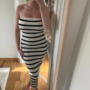 Randig axelbandslös långklänning - Säljer en ribbad, axelbandslös långklänning med svartvita horisontella ränder. Klänningen är tight och figurnära med stretchigt material som sitter snyggt på kroppen. Perfekt för dig som gillar en stilren och trendig look.