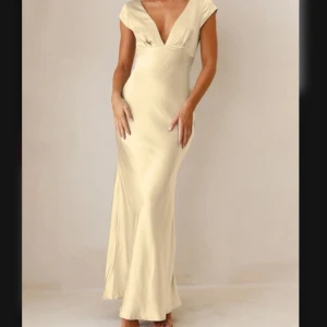 Ljusgul balklänning i satin  - Supersnygg långklänning i champagnefärgad satin med djup v-ringning och korta ärmar. Klänningen har en öppen rygg med twistad detalj och figurnära passform som ger en elegant siluett. Perfekt för dig som vill sticka ut med en lyxig look. Balklänning i satin. Helt oanvänd. Köpt för ca 2000kr