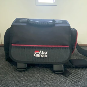 Svart axelväska Abu Garcia med röda detaljer - Rymlig svart axelväska från Abu Garcia med röda detaljer och vit logga. Väskan har robust handtag, justerbar axelrem och flera fack, perfekt för att organisera småprylar. Tillverkad i slitstark polyester och har dragkedjor för enkel åtkomst. Rostig på vissa delar, perfect för någon som vill börja fiska. Det är 3 lådor i den och massor av fickor.