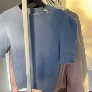 Ljusblå stickad tröja från Zara - Superfin ljusblå stickad tröja från Zara med korta ärmar och glittriga dekorationsknappar vid axeln och ärmslutet. Tröjan har ribbade muddar och en klassisk rund halsringning. Tröjan är som ny och i storlek M, dock ganska liten i storleken