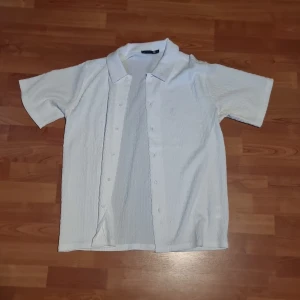 Vit kortärmad skjorta/ overshirt med struktur - Säljer en vit skjorta/ overshirt med korta ärmar och krage. Skjortan har en lätt strukturerad yta och knappar framtill. Perfekt för varma dagar och enkel att matcha med jeans eller shorts.