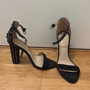 Svarta sandaler med nitar från Trafaluc - Svarta sandaler med bred klack och öppen tå från Trafaluc. Skorna har remmar med silverfärgade nitar och spänne runt vristen. Klacken och hälen är dekorerade med både runda och spetsiga nitar. Yttermaterial i mocka-look.