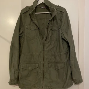 Grön fieldjacket - Snygg olivgrön field jacka med fyra stora fickor framtill och dragkedja. Jackan har hög krage, axelklaffar. Perfekt för lager på lager och nu under sommaren! Skick 9/10.