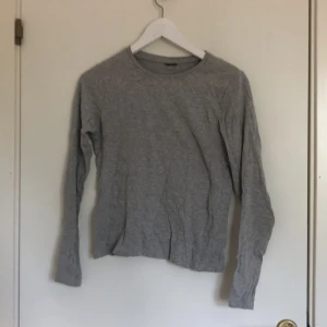 Grå basic långärmad tröja - Enkel och stilren grå långärmad tröja i mjuk bomull. Tröjan har rund halsringning och en rak passform som funkar till det mesta. Perfekt att ha som basplagg i garderoben och lätt att matcha med andra kläder.