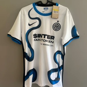 Inter Milan vit bortatröja Nike - Inter Milans officiella bortatröja från Nike i vitt med blå och svarta detaljer. Tröjan har ett unikt ormmönster, svart Inter-logga och sponsortryck på bröstet. Kortärmad modell i lätt och ventilerande material, perfekt för match eller träning.