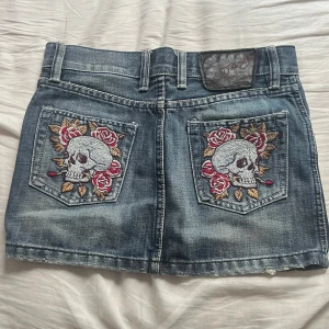 Ed Hardy jeanskjol med döskallebrodyr - Snygg jeanskjol från Ed Hardy med broderade döskallar och rosor på bakfickorna. Klassisk blå denim med slitningar och dragkedja framtill. Trendig minikjol, det står storlek S men skulle säga att den passar en XS/XXS. Det är bara att skriva om ni har frågor💞💞💞