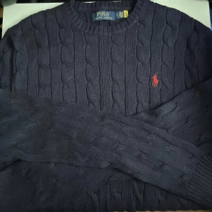 Mörkblå stickad tröja Polo Ralph Lauren - Lite smutsig för den har bara legat i garderoben,   Kabelstickad mörkblå tröja från Polo Ralph Lauren med klassisk röd broderad logga på bröstet. Tröjan har rund halsringning och långa ärmar, perfekt för lager-på-lager-stil. Materialet är mjukt och skönt stickat. Skriv om du är intresserad eller vill ha mer bilder