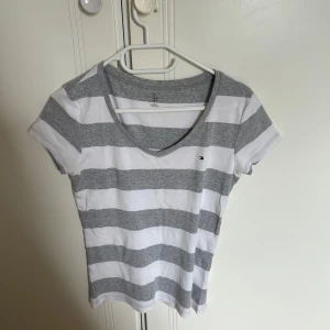 Randig t-shirt från Tommy Hilfiger - Snygg t-shirt från Tommy Hilfiger med breda grå och vita ränder. T-shirten har v-ringning, korta ärmar och en liten broderad logga på bröstet. Tillverkad i mjuk bomull som känns skön mot huden. Perfekt för en avslappnad och stilren look.
