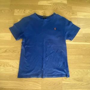 Blå t-shirt från Polo Ralph Lauren - Klassisk blå t-shirt från Polo Ralph Lauren med liten broderad logga i orange på bröstet. T-shirten har rund halsringning och är tillverkad i mjuk bomull. Perfekt basic-plagg med snygg passform och stilren look. Priset kan diskuteras.
