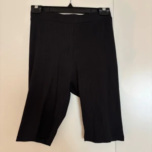 Svarta ribbade cykelshorts Lager 157 - Svarta cykelshorts från Laget 157 i ribbat material. De har en midwaist passform och är stretchiga och bekväma, perfekta för en sportig look eller till oversized t-shirt. Klassisk svart färg och enkel design som funkar till det mesta.