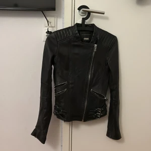 Svart skinnjacka Trafaluc XS - Snygg svart skinnjacka från Trafaluc i äkta läder. Jackan har coola detaljer som sned dragkedja, quiltade axlar, dragkedjor vid ärmslut och dubbla spännen i midjan. Perfekt för dig som vill ha en edgy look med stilren passform.