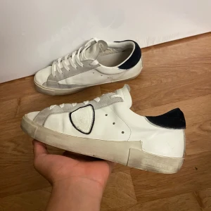 Vita sneakers från Philippe Model - Snygga vita sneakers från Philippe Model med grå mockadetaljer och svart häl. Klassisk låg modell med snörning och rund tå. Skorna har en vit sula och den ikoniska sköld-loggan på sidan. Perfekta för en clean och stilren look.