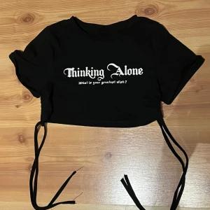 Svart croppad topp med snörning - Cool svart croppad topp med vit text 'Thinking Alone' framtill. Toppen har korta ärmar och snörning i sidorna som ger en snygg detalj och möjlighet att justera passformen. Perfekt för dig som gillar edgy streetstyle.