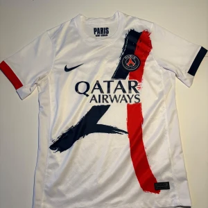 PSG vit Nike fotbollströja Dri-FIT - Paris Saint-Germain fotbollströja i vitt med stora penseldrag i mörkblått och rött framtill. Broderat PSG-märke på bröstet, Nike-logga och Qatar Airways-tryck. Tillverkad i lätt, ventilerande Dri-FIT-material. Kortärmad modell med rund hals.