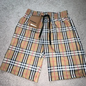Rutig Burberry shorts med snörning - Snygga shorts från Burberry i klassiskt beige, svart, vit och röd rutig design. De har elastisk midja med svart snörning och är tillverkade i ett lätt och bekvämt material, perfekt för sommardagar. Ikonisk look som sticker ut och passar till streetwear.