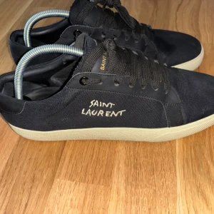 Svarta Saint Laurent sneakers - Snygga svarta sneakers från Saint Laurent med vit broderad logga på sidan och svart läderdetalj vid hälen. Ovandel i canvas och klassisk vit sula. Skorna har svarta snören och en stilren, minimalistisk look som passar till allt.