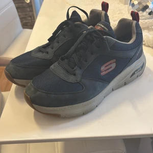 Mörkblå Skechers Arch Fit sneakers - Snygga mörkblå Skechers Arch Fit sneakers med röd logga på sidan. Skorna är vattentäta och har en robust, ljusgrå sula. Ovandelen är i mesh och mockaliknande material, med svarta snören och bekväm vadderad insida. Perfekta för dig som vill ha både stil och komfort.