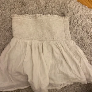 tubtopp från Gina Tricot - Supersöt vit bandeau tubtopp från Gina Tricot med smockad överdel och volangig nederdel. Toppen är axelbandslös och har en luftig känsla, perfekt för varma dagar. Enkel att matcha med jeans eller kjol för en chill look.