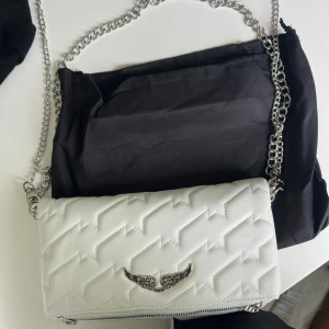 Vit quiltad axelväska Zadig & Voltaire - Snygg vit axelväska från Zadig & Voltaire i quiltat skinn med coolt blixtmönster. Väskan har silverfärgad kedjerem, dragkedja upptill och ikonisk vingdetalj framtill. Perfekt storlek för mobilen och det viktigaste. Kommer med dustbag.