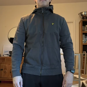 Mörkgrå hoodiejacka från Lyle & Scott - Snygg mörkgrå hoodiejacka från Lyle & Scott med gul broderad logga på bröstet. Jackan har dragkedja hela vägen, huva med snörning och två fickor framtill. Perfekt för dig som gillar en clean och sportig stil.