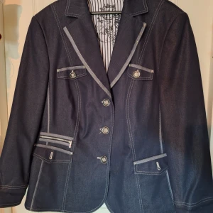 Mörkblå jeanskavaj från Gerry Weber - Snygg mörkblå jeanskavaj från Gerry Weber med markerade sömmar, dekorativa knappar och coola detaljer som dragkedja på ena fickan. Klassisk krage, långärmad och randig på insidan.