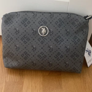 Shoppingväska från U.S. Polo Assn. - Snygg shoppingväska från U.S. Polo Assn. i mörkgrått med svart mönster och logga. Väskan har dragkedja upptill, silverfärgade metalldetaljer och ett diskret all-over print med både logotyp och polospelare. Tillverkad i syntetmaterial och har en praktisk form. Helt ny aldrig använd. Nytt pris 1200kr 