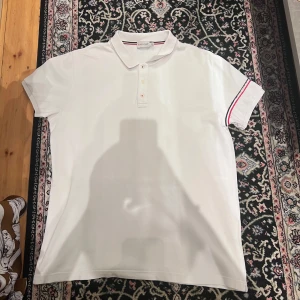 Vit pikétröja från Moncler XL - Vit pikétröja från Moncler i klassisk modell med krage och knappar framtill. Ärmarna har en snygg detalj med röd och marinblå rand. Tröjan är tillverkad i bomull och har en clean, sportig vibe som passar perfekt till jeans eller shorts.