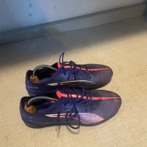 Blå och lila Puma fotbollsskor - Säljer ett par snygga fotbollsskor från Puma i mörkblått med rosa och vita detaljer. Skorna har snörning och en lätt, syntetisk ovandel för bästa känsla på planen. Sulan har fasta dobbar i rosa och blått för bra grepp på gräs.