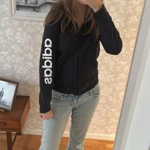 Svart Adidas hoodie med dragkedja - Svart hoodie från Adidas med vit logga längs ena ärmen. Tröjan har huva med snörning, dragkedja framtill och två fickor. Skön passform och tillverkad i mjuk bomullsmix, perfekt för chill eller träning.