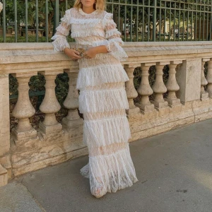 Vit spetsklänning med volanger - Drömmig långklänning i vit spets med flera lager volanger och utsvängda ärmar. Klänningen har en romantisk känsla med transparent spets och rund halsringning. Perfekt för dig som vill sticka ut med en unik och elegant look. Bröllopsklänning carmine dress