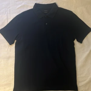 Mörkblå pikétröja från Massimo Dutti - Snygg mörkblå pikétröja från Massimo Dutti med klassisk krage och knappslå framtill. Tröjan har korta ärmar och en stilren, enkel design som passar perfekt till jeans eller chinos. Tillverkad i 100% bomull och är i storlek M