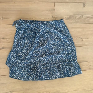 Blå blommig omlottkjol med volang - Supersöt blå kortkjol med vitt blommmönster. Använd ett fåtal gånger, så jättefint skick!!🩷☀️