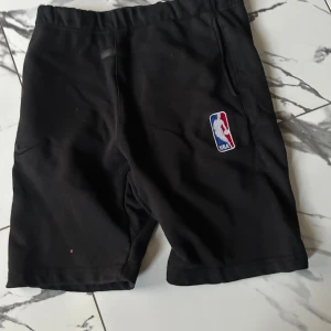 Svarta NBA shorts Fear of God Essentials - Svarta shorts från Fear of God Essentials med broderad NBA-logga på ena benet. De har elastisk midja och sidofickor, perfekt för basket eller chill. Mjukt material som känns skönt mot huden och en clean streetwear-look.