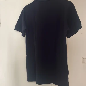 Svart pikétröja Polo Ralph Lauren S - Klassisk svart pikétröja från Polo Ralph Lauren i modellen Custom Slim Fit. Tröjan har korta ärmar, krage med två knappar och den ikoniska röda loggan broderad på bröstet. Perfekt för en stilren och sportig look.