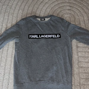 Grå sweatshirt från Karl Lagerfeld - Snygg grå sweatshirt från Karl Lagerfeld med stort vitt logotryck på bröstet. Tröjan har rund halsringning och långa ärmar, perfekt för en clean och stilren look. Mjuk bomullskänsla och klassisk passform. Storlek M!😊