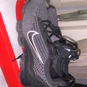 Nike Vapormax grå sneakers - Snygga Nike Vapormax sneakers i grått och svart med stickad ovandel och tydlig vit swoosh på sidan. Skorna har svart snörning och genomskinlig sula med luftbubblor för extra komfort. Perfekta för dig som gillar sportig och modern stil.