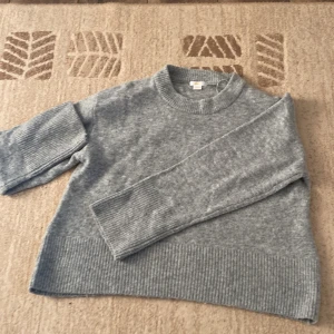 Grå stickad tröja från Gina Tricot - Mysig grå stickad tröja från Gina Tricot med breda ribbade muddar vid ärmslut och nederkant. Tröjan har en klassisk rund halsringning och lång ärm, perfekt för lager-på-lager under kyliga dagar. Enkel och stilren design som funkar till allt. Riktigt fint skick då den är använd ett fåtal gånger!