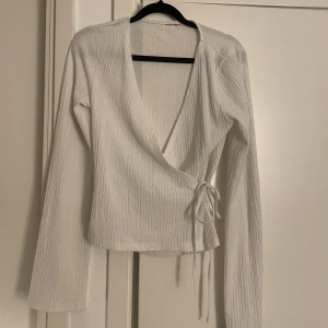 Vit omlottopp med knytband - Säljer en vit omlottopp med långa ärmar och knytband i sidan. Toppen har en djup v-ringning och är tillverkad i ett lätt, strukturerat tyg som ger en snygg och modern look.