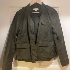 Olivgrön utilityjacka med fickor - Säljer en olivgrön utilityjacka från H&M med flera stora fickor framtill och axelklaffar. Jackan har lång ärm, rak passform och stängs med dolda tryckknappar. Perfekt för lager-på-lager under höst och vår. En av knapparna är trasig, men syns inte när den är stängd.