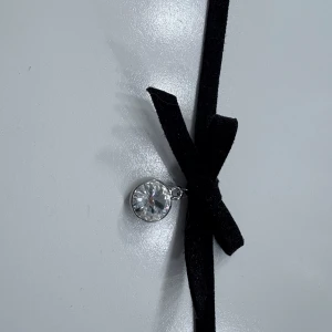 Choker med svart band och glittrande sten - Snygg choker med ett smalt svart band i sammet och en glittrande rund sten i silverinfattning. Bandet knyts i en rosett framtill för en extra detalj. Perfekt för att lyfta din outfit.