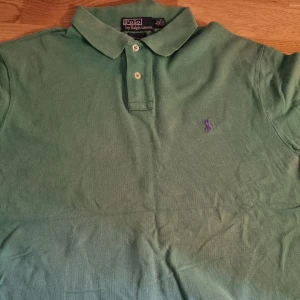 Grön pikétröja från Polo Ralph Lauren - Snygg grön pikétröja från Polo Ralph Lauren med klassisk krage och två knappar framtill. Liten lila broderad logga på bröstet. Perfekt till en avslappnad men stilren look.