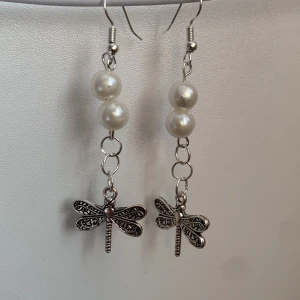 Silverfärgade örhängen med pärlor och trollslända - Unika örhängen med två vita pärlor och en detaljrik trollslända i silverfärg. Hängande modell som ger ett lekfullt och stilrent intryck. Perfekta för dig som vill ha något annorlunda och snyggt till din outfit.
