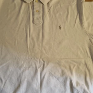 Vit pikétröja från Ralph Lauren - Klassisk vit pikétröja från Ralph Lauren med korta ärmar och broderad logga på bröstet. Tröjan har krage och knäppning med två knappar framtill. Perfekt för en stilren och sportig look.