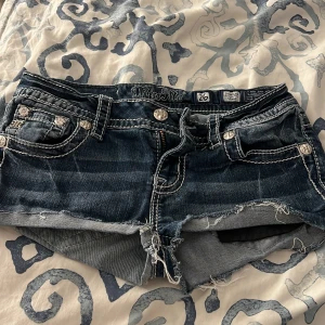 Jeansshorts med vingar från Miss Me - Säljer ett par blå jeansshorts från Miss Me med slitna kanter och snygga broderade vingar i silver på bakfickorna. Shortsen har låg midja, dekorativa nitar och detaljerade sömmar. Perfekta för sommaren! Av klippta från miss me jeans🩷