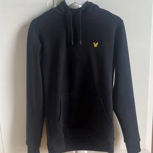 Svart hoodie från Lyle & Scott - Svart hoodie med huva och dragsko från Lyle & Scott. Tröjan har en gul broderad logga på bröstet och en stor magficka framtill. Perfekt för en avslappnad stil.